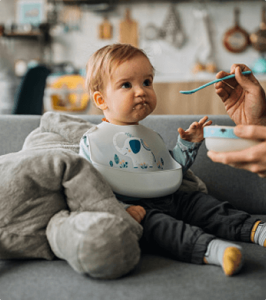 Baby being spoon fed on a couch.